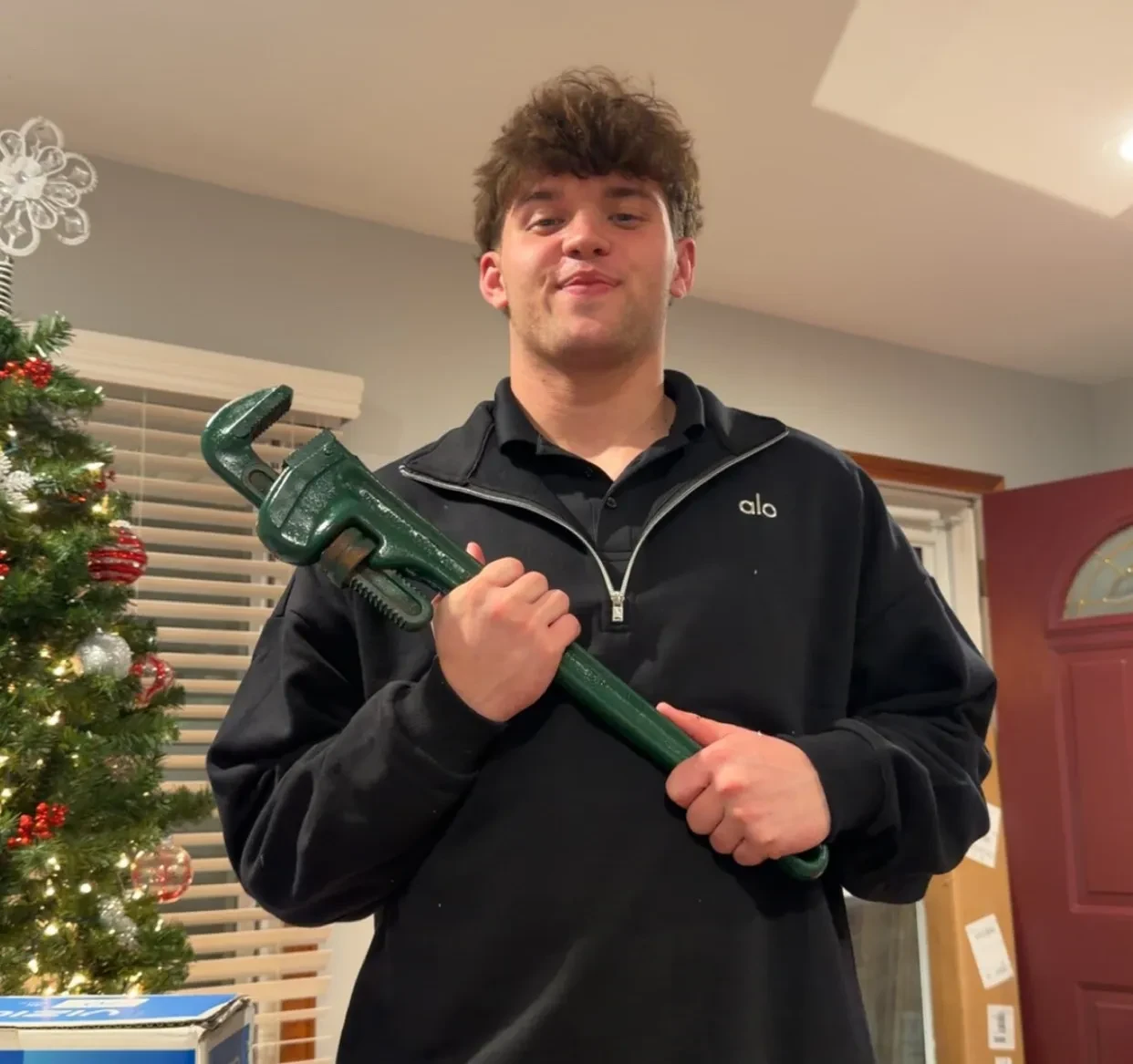 Noah Knigga in a black quarter zip holding a green pipe wrench